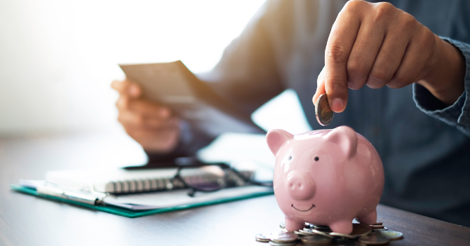 Man putting a coin into a piggy bank on a desk.